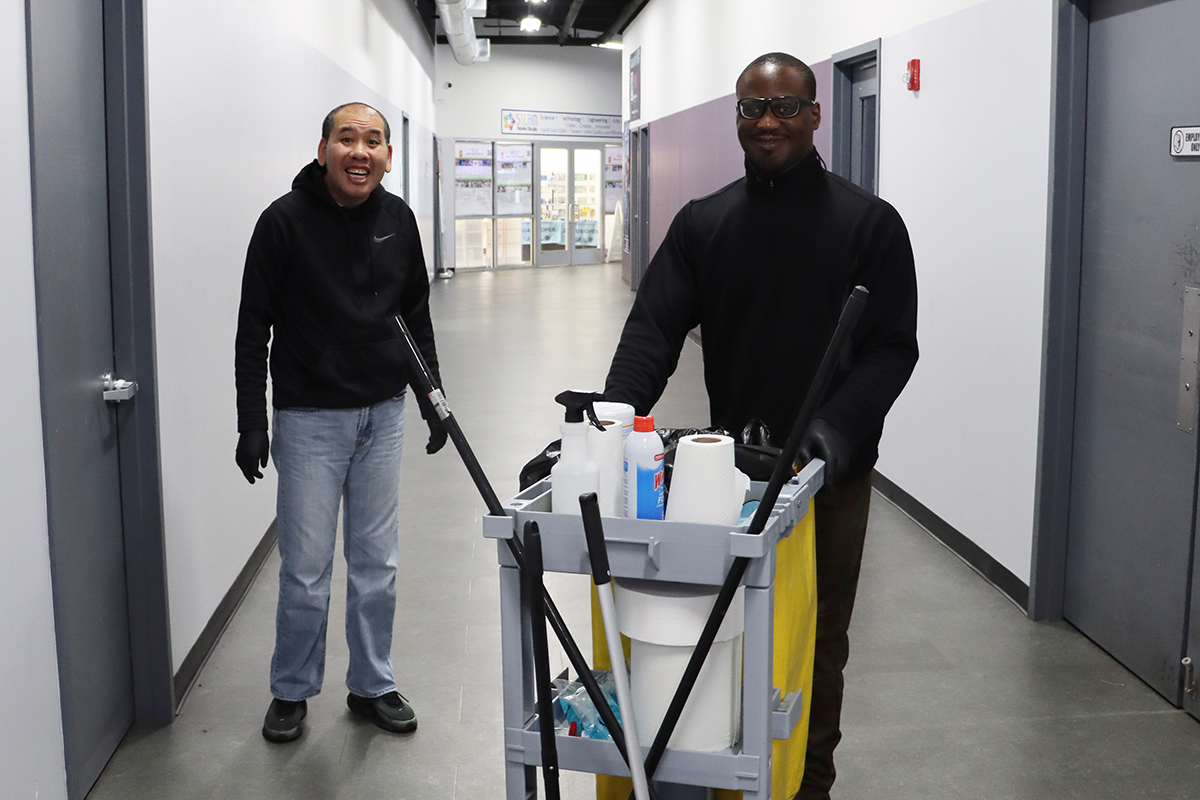 Two men smile at work in front of their cleaning cart.